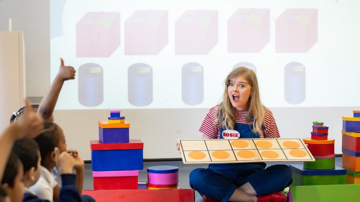 A learning facilitator is sitting on the floor surrounded by children. She holds a game and looks at them with a surprise face.