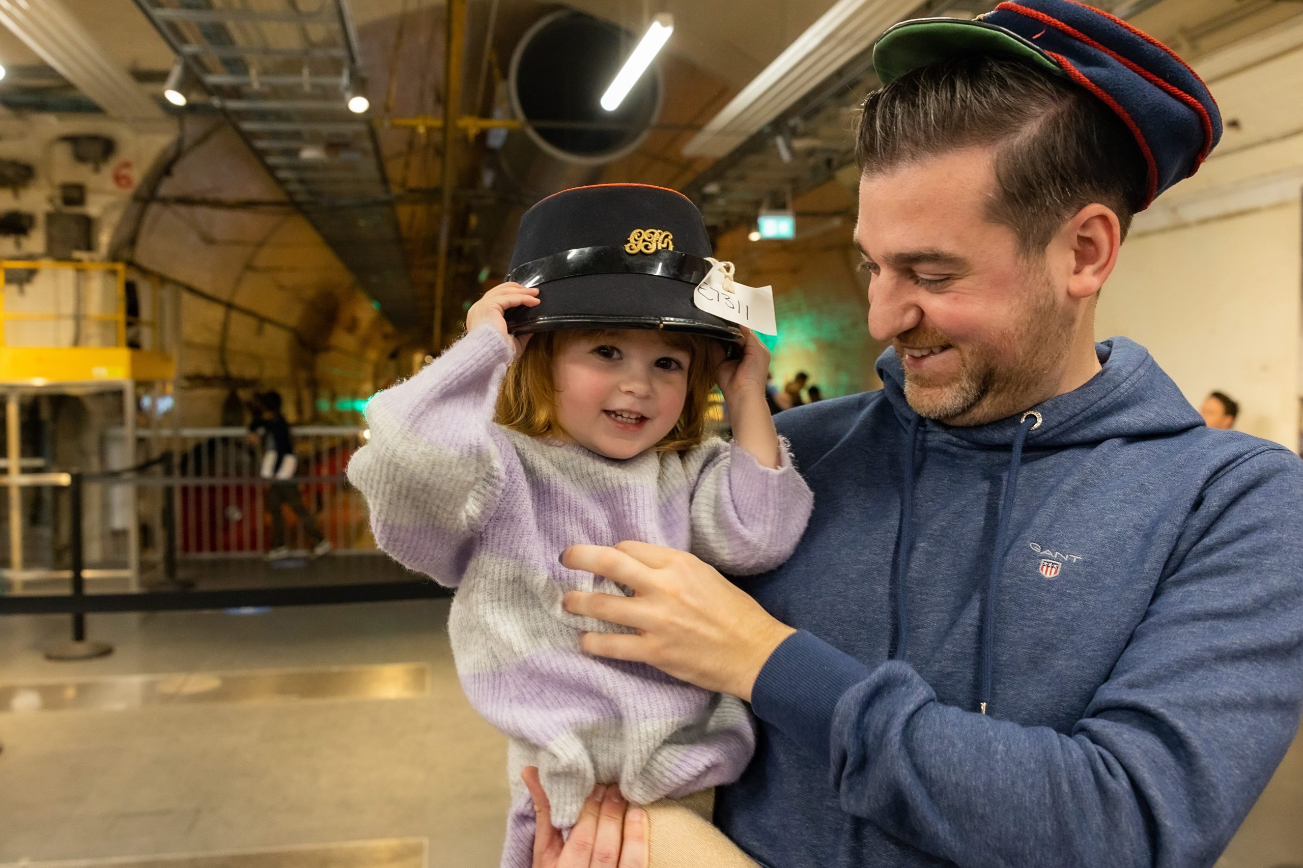 A happy dad is carrying his little girl, she is wearing an old Postie hat, which is way too big for her.