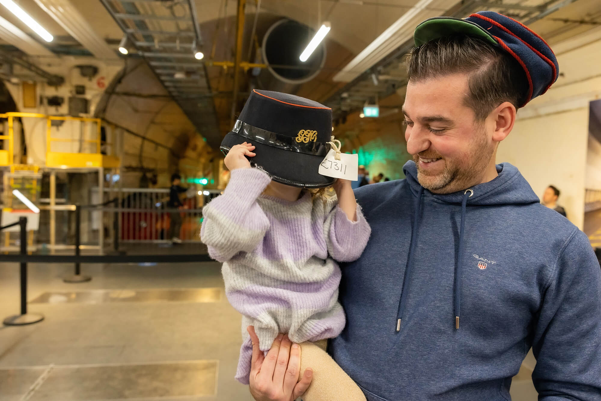 A man, with dark hair and light skin holds a little girl with blonde hair and light skin. She is playing dress-up with a hat which is far too big for her. The man is laughing and looking at her fondly.