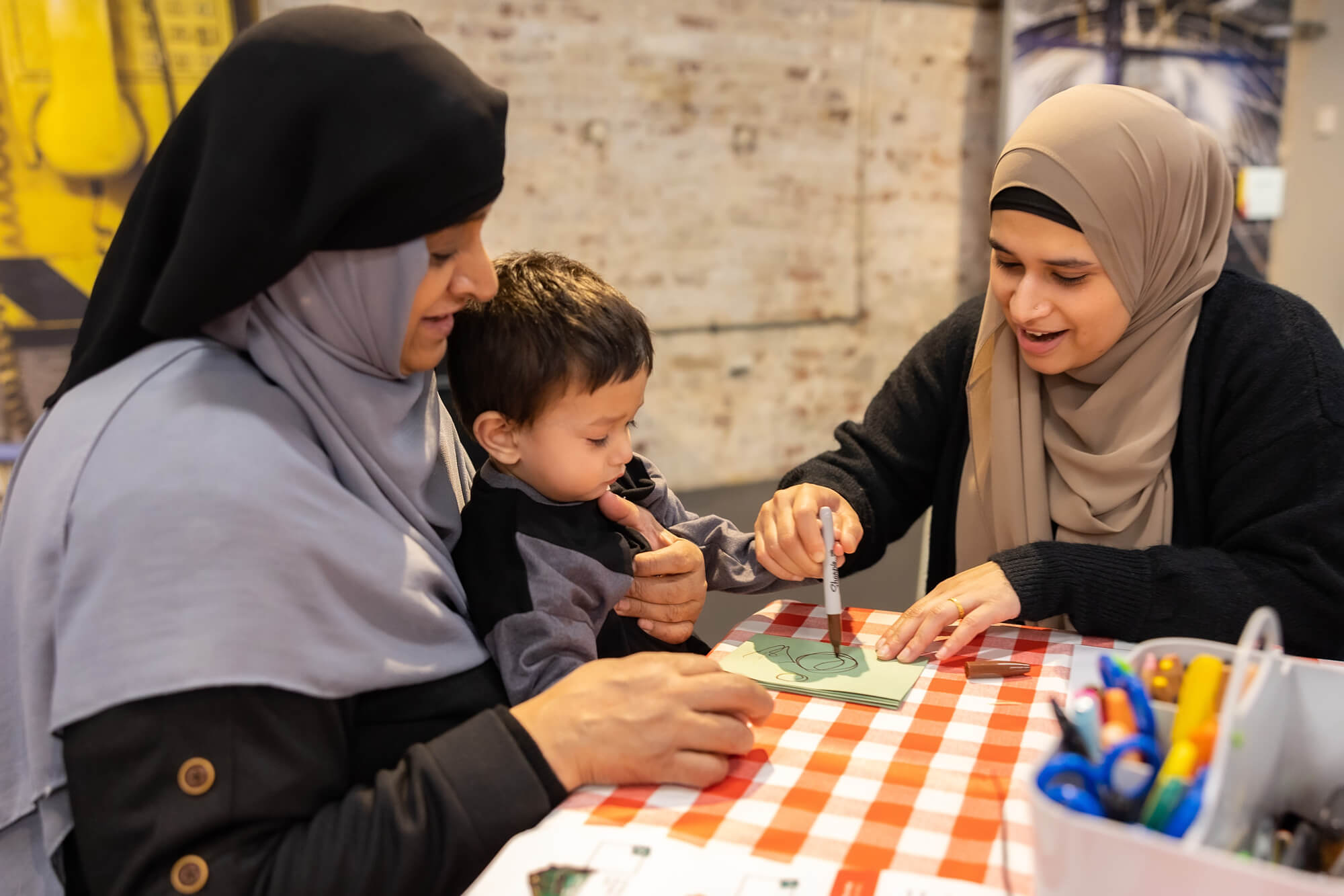Two women wearing Hijabs are helping a little boy with his arts and crafts project. The dark haired boy sits on the women on the right's lap.