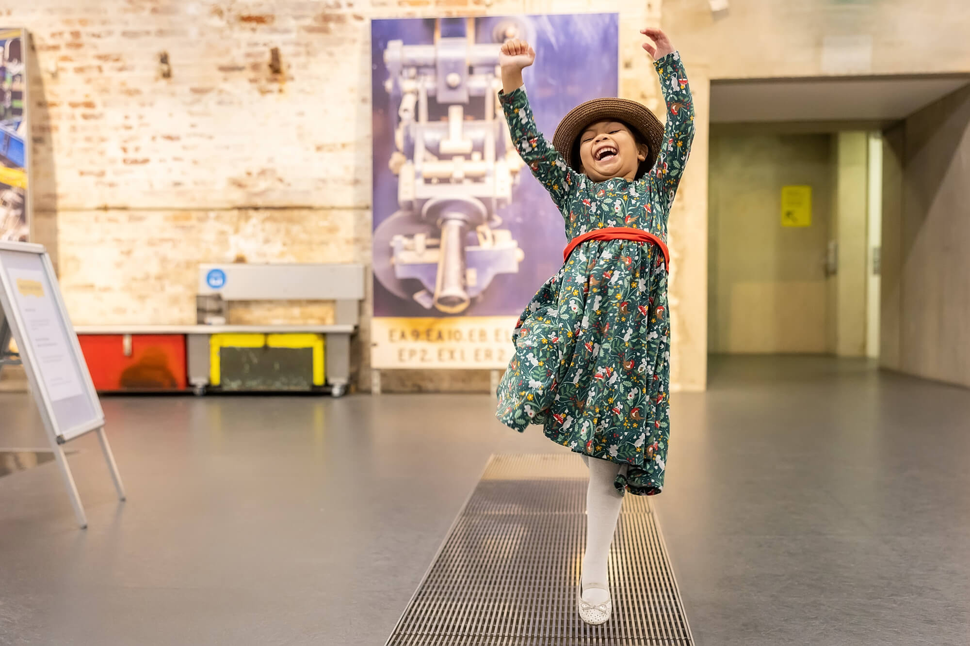 A little girl with brown skin and dark hair jumps for joy. Her arms are in the air and her mouth is open as if laughing. She is playing dress up, wearing an old straw hat.
