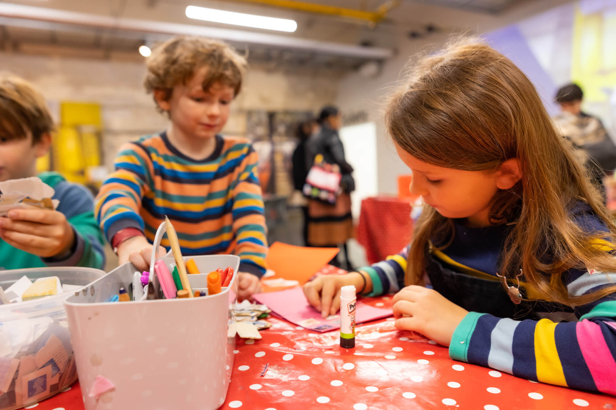 Three brown-haired children wearing long-sleeved t-shirts taking part in an arts and crafts activity. They are holding Pritt Sticks and pencils.